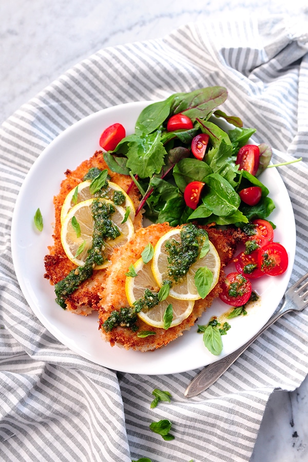 Breaded asiago chicken topped with lemon wheels and pesto served on a plate with a leafy green salad